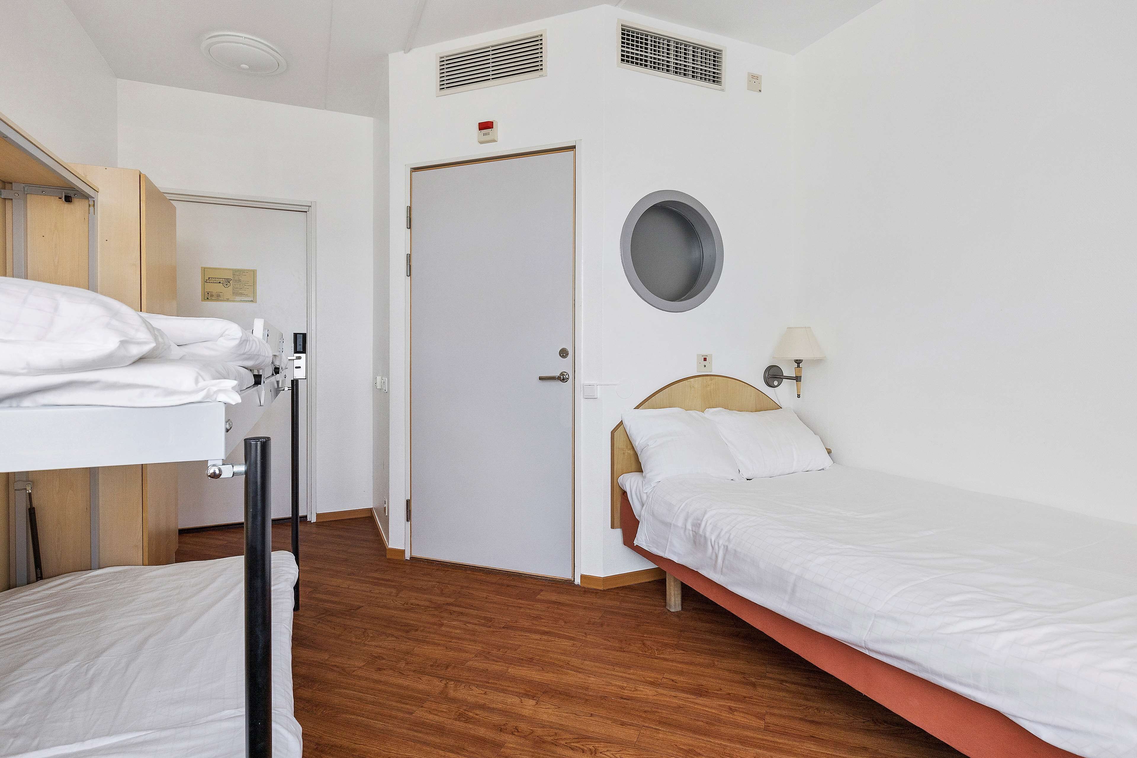 A hotel room with a bunk bed, a single bed, a round mirror, a lamp, and a door with a sign.