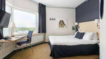 A hotel room with a bed, a desk with a chair, a window with curtains, a wall-mounted TV, and a painting.