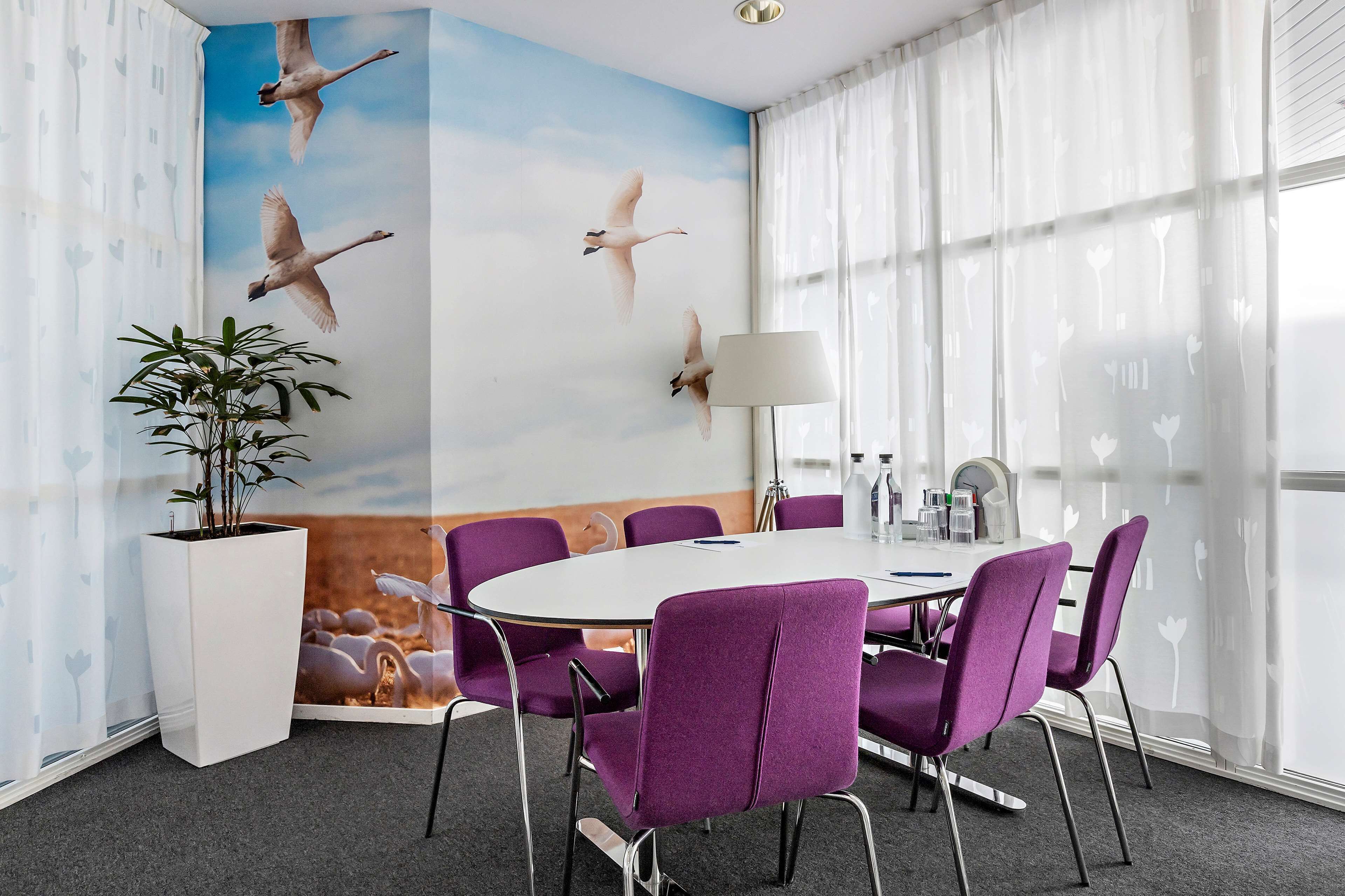 Un salón de conferencias con una mesa redonda, sillas moradas, una lámpara blanca y un mural con pájaros volando.