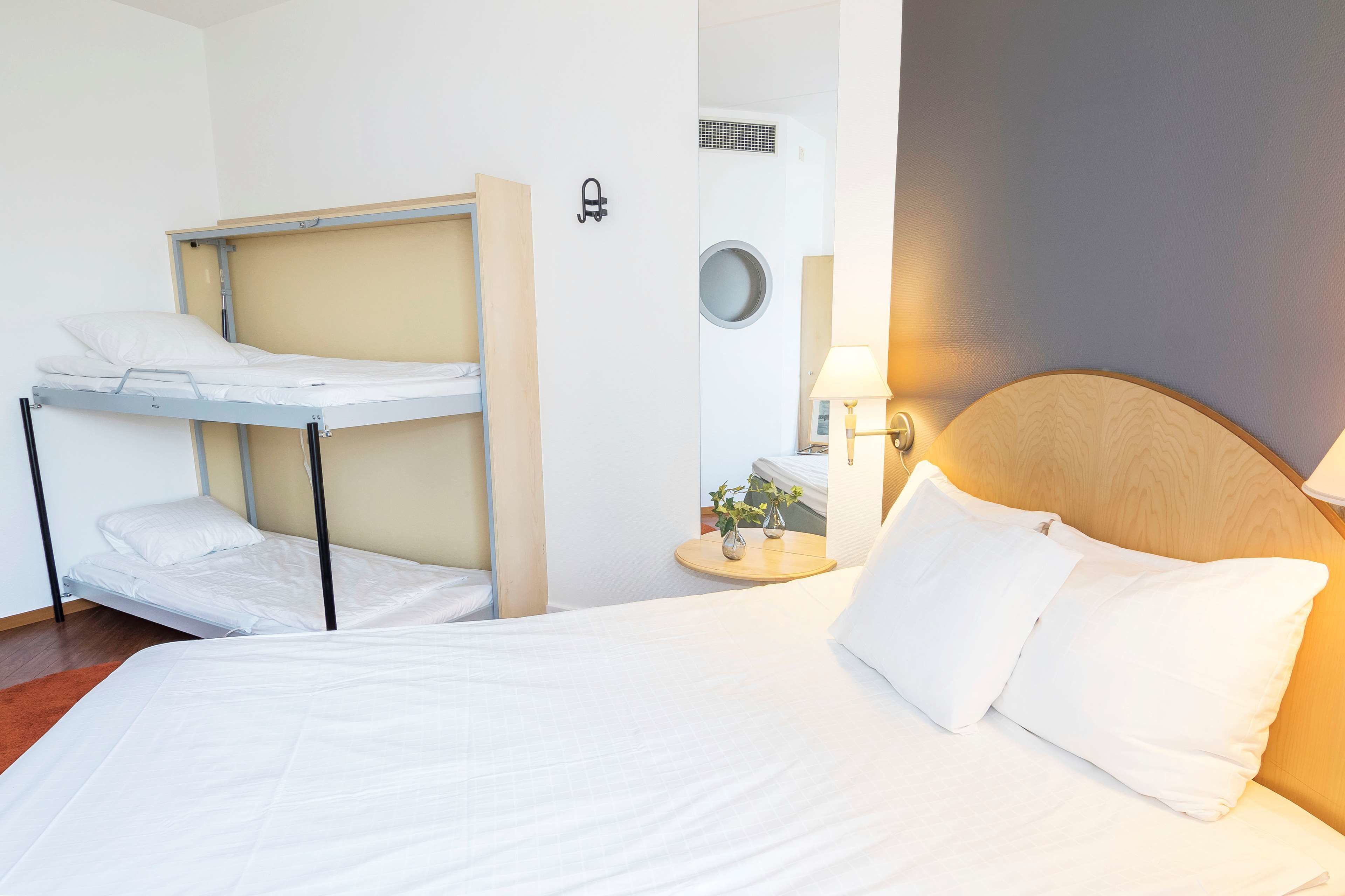 Hotellihuone, jossa on yksi sänky, kerrossänky, yöpöytä lampun kanssa ja pieni pyöreä peili.