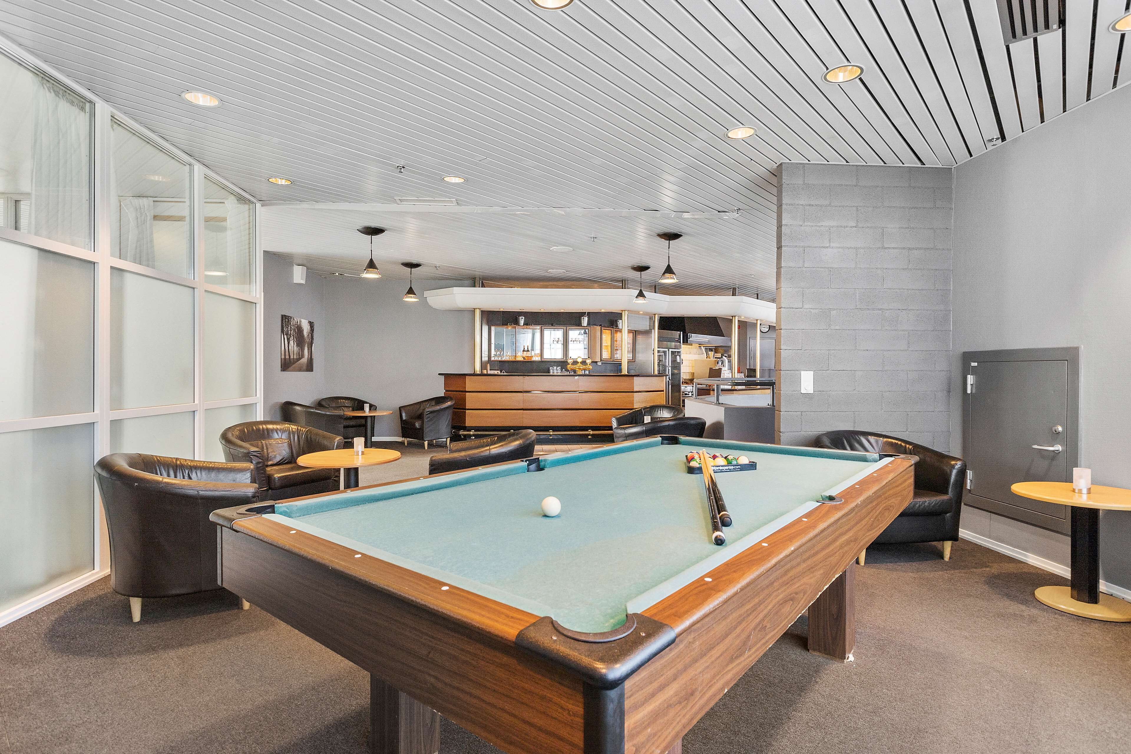 Une table de billard dans une pièce comprenant un bar, des fauteuils en cuir et de grandes fenêtres offrant une vue dégagée.