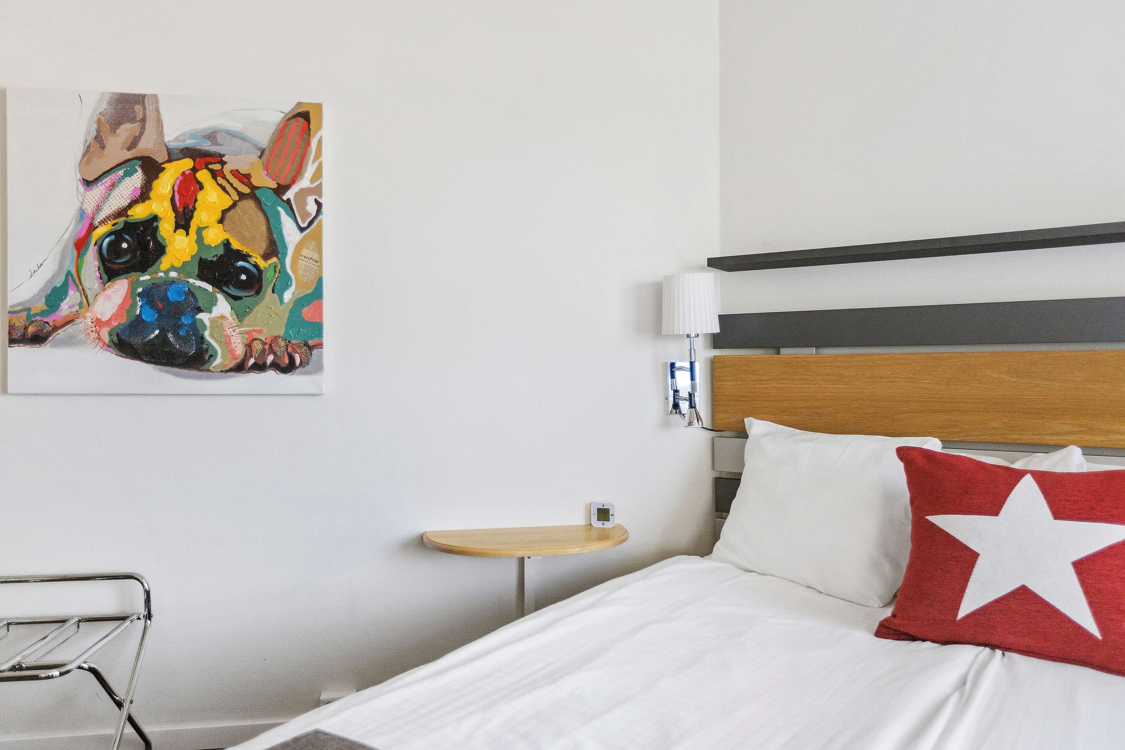 Quarto de hotel com cama, mesa de cabeceira, abajur e uma obra de arte na parede retratando um cão.
