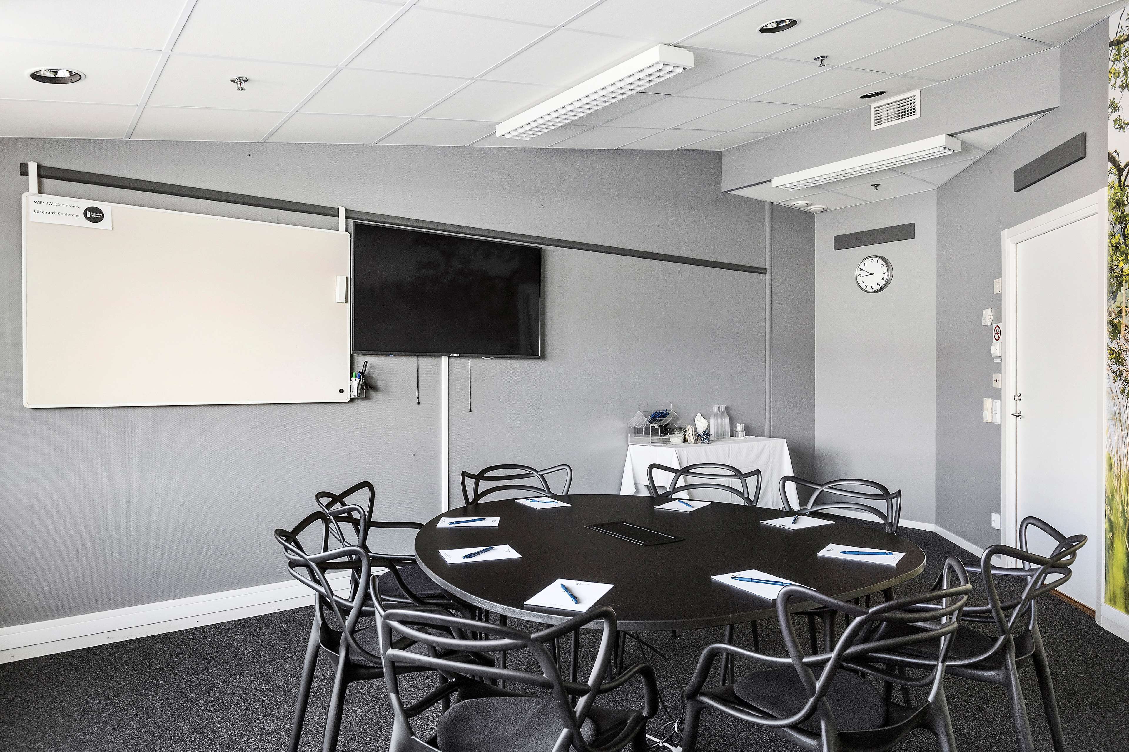 Ett konferensrum med ett runt bord, åtta stolar, en whiteboard, en platt-TV och en väggklocka.