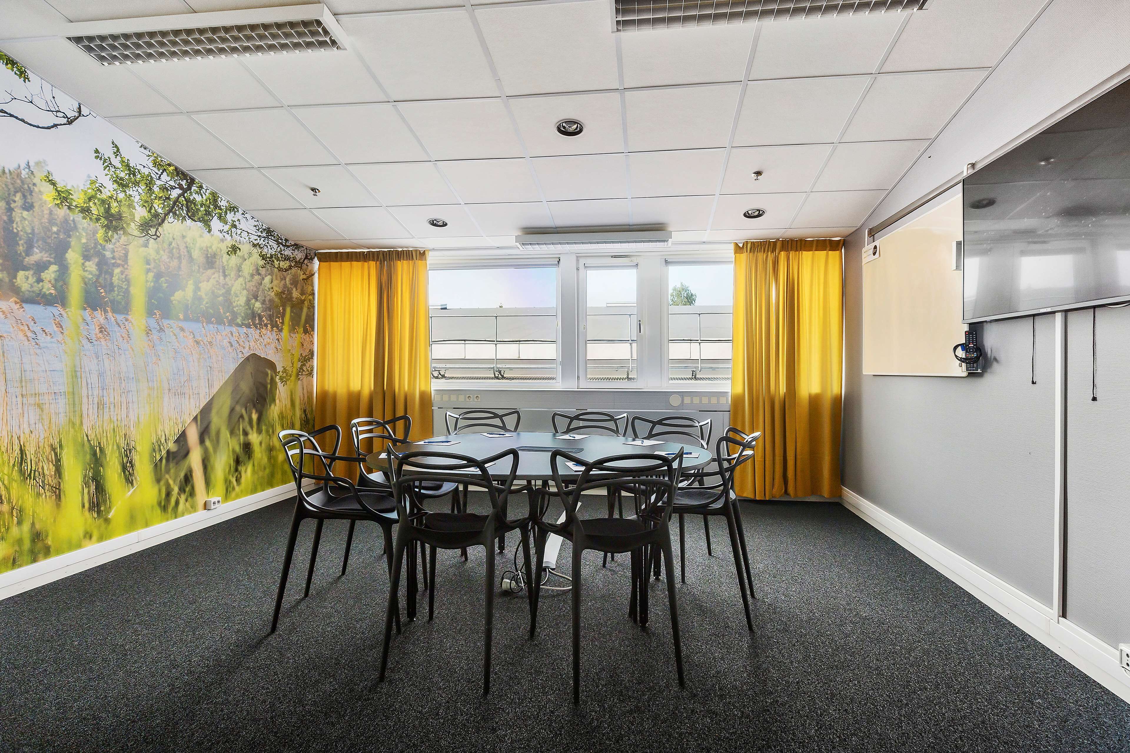 Un salón de conferencias con una mesa redonda, sillas negras, un ventanal con cortinas amarillas y un mural de paisajes naturales en la pared.