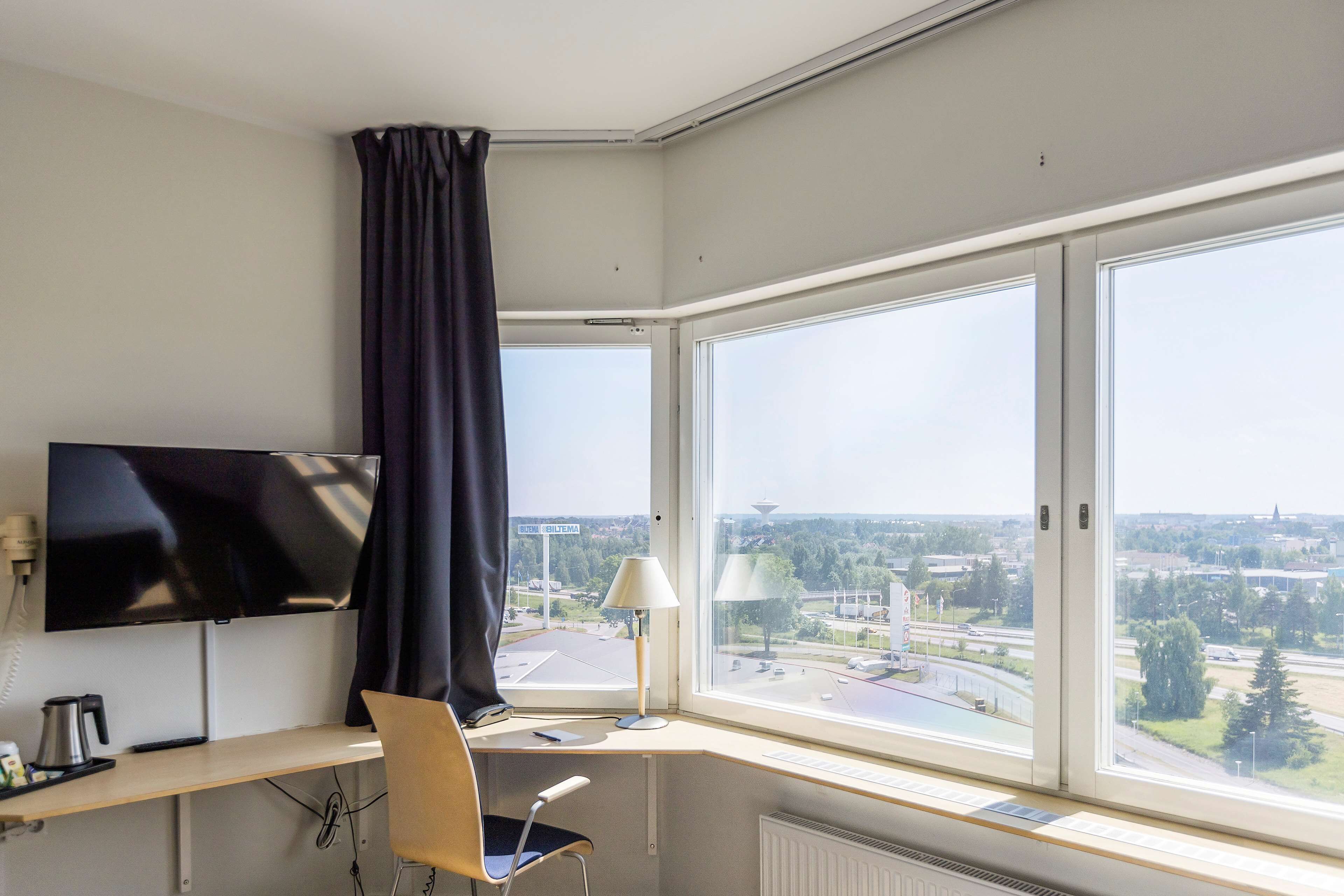 Una habitación de hotel con un ventanal grande, un escritorio con ordenador, una silla, un televisor y una lámpara.