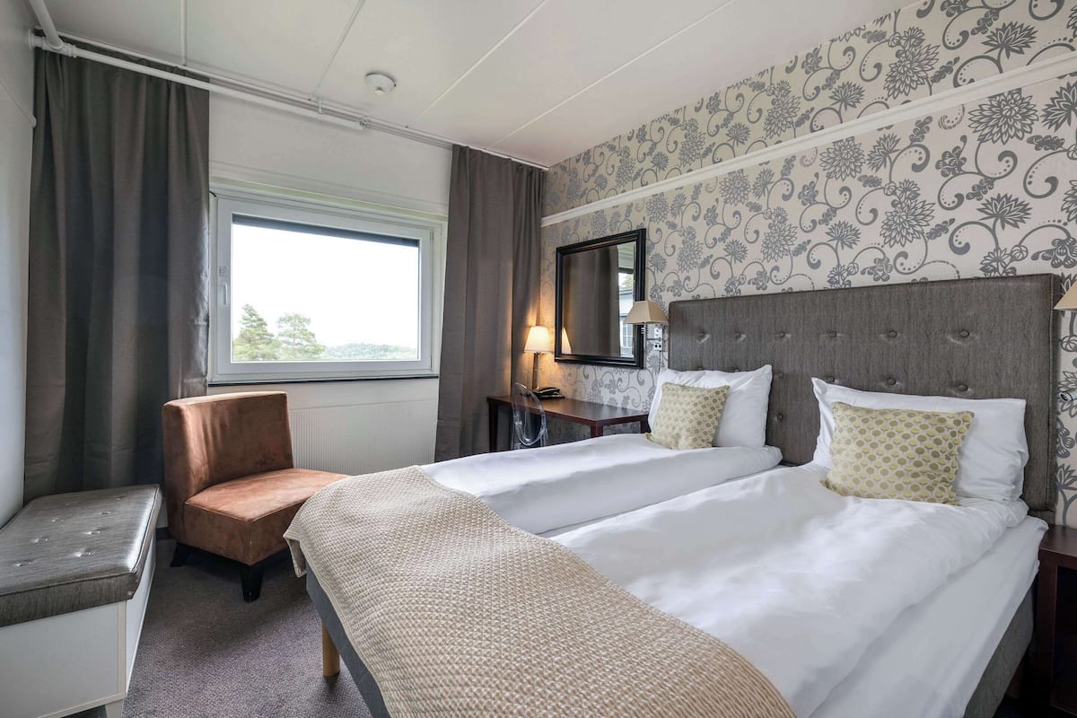 A hotel room with two beds, a desk, a chair, a window with curtains, and a patterned wallpaper.