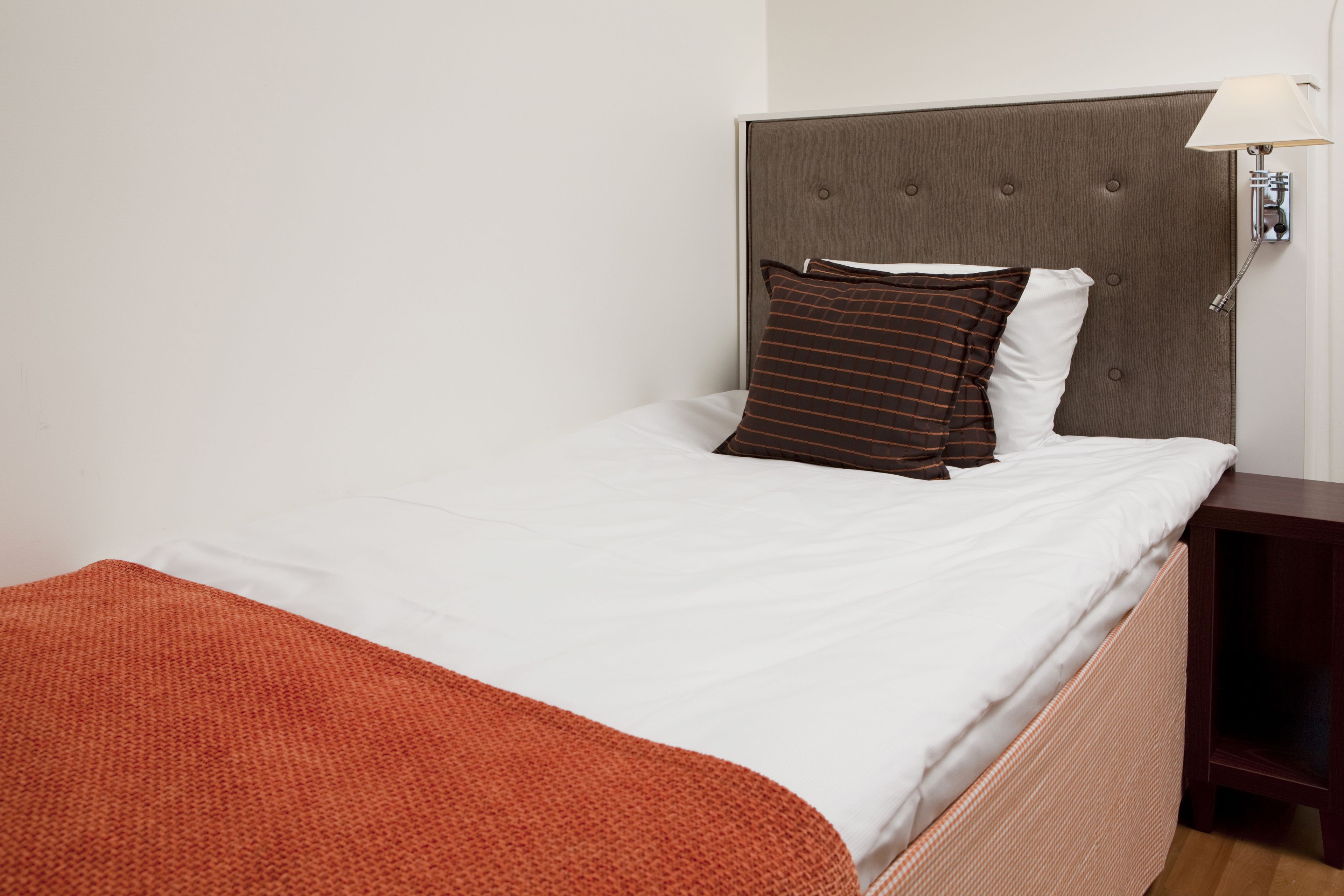 Una cama bien hecha con un cojín a rayas marrones y uno blanco, una mesita de noche de madera con una lámpara, y una manta roja sobre la cama.