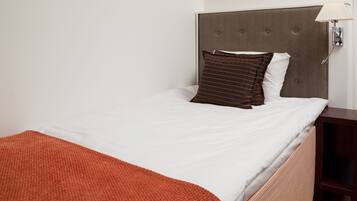 Una cama bien hecha con un cojín a rayas marrones y uno blanco, una mesita de noche de madera con una lámpara, y una manta roja sobre la cama.