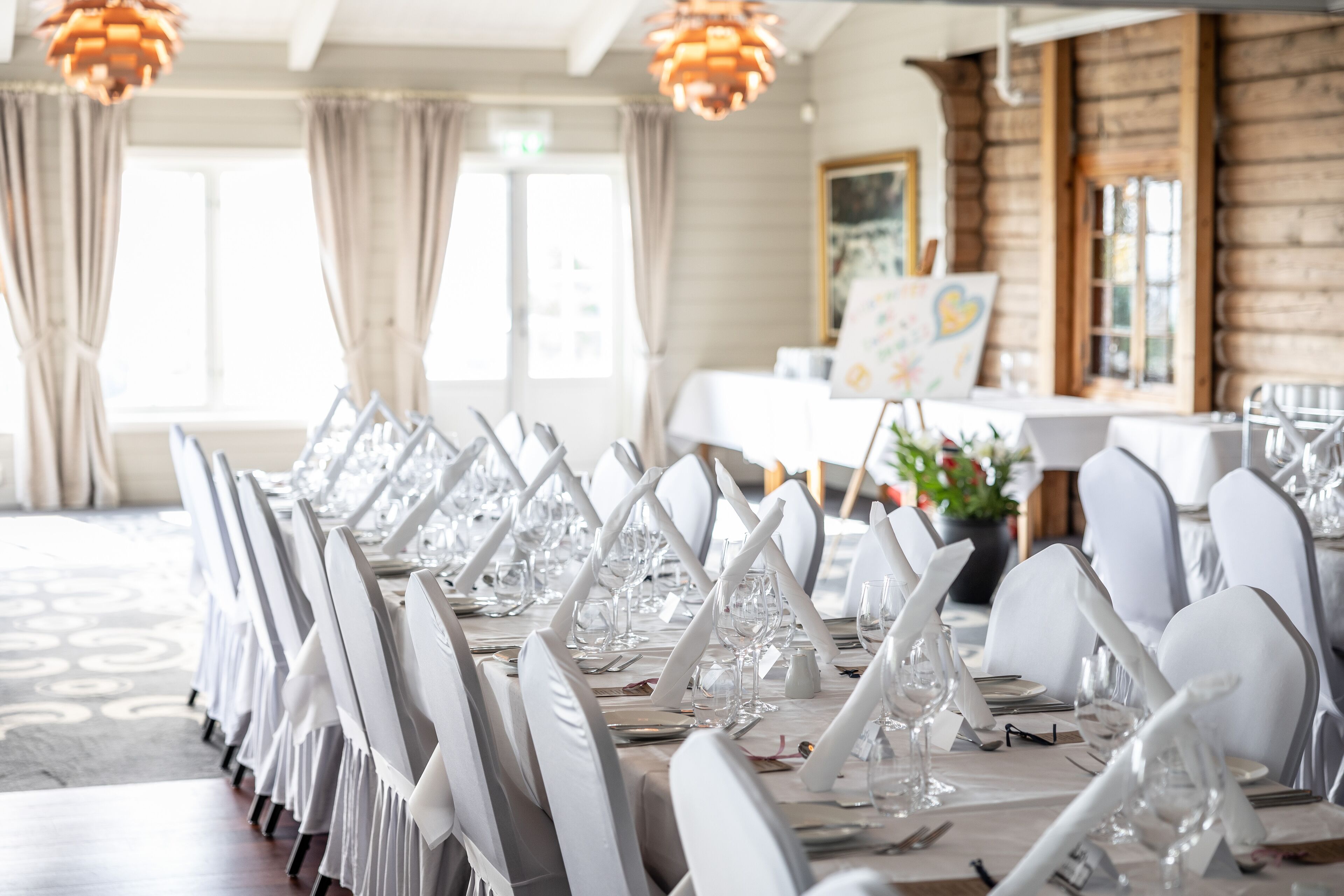 Un área de comedor con sillas y mesas blancas dispuestas para un evento formal, con una pared rústica de madera y ventanas amplias.