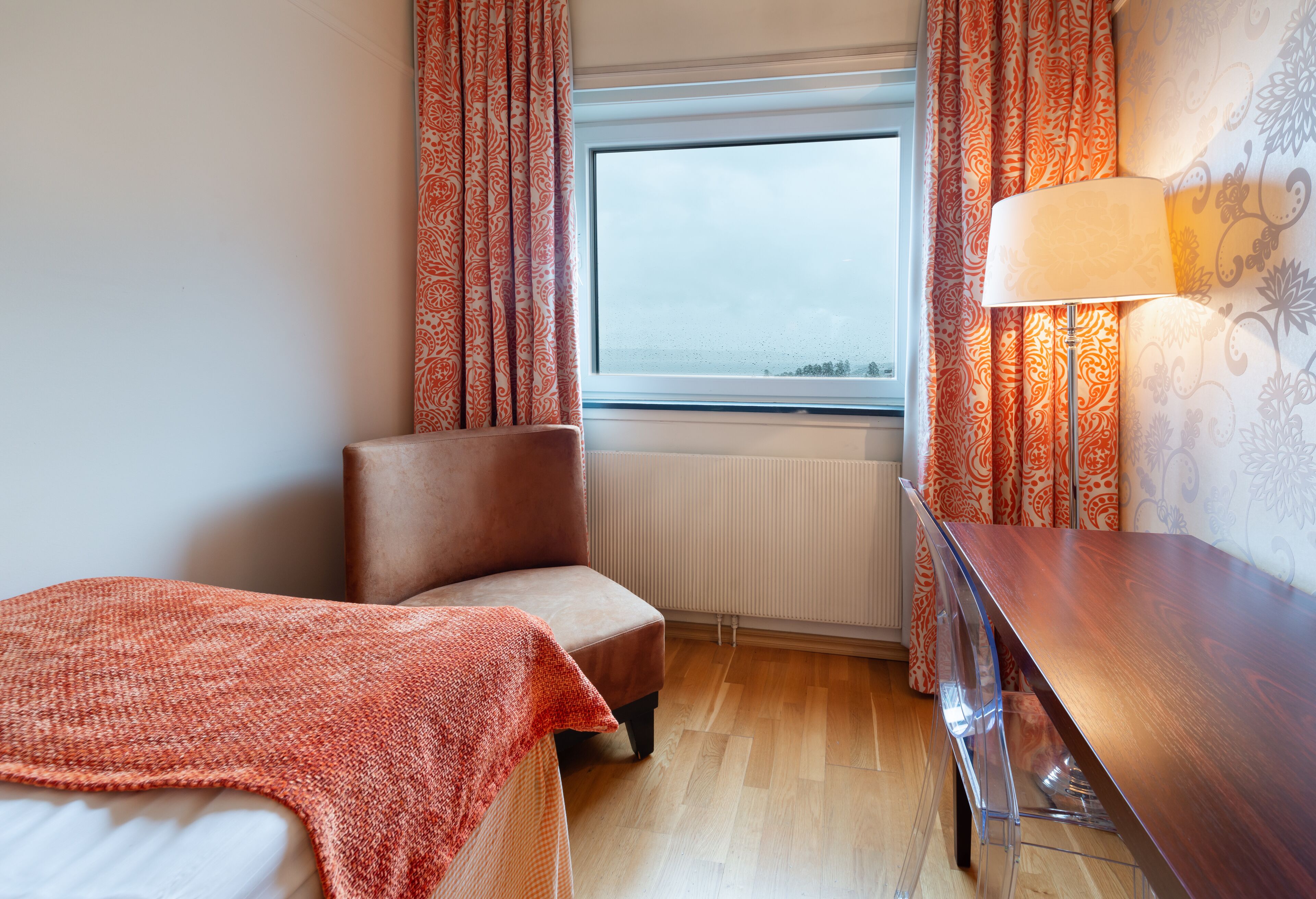 Una habitación de hotel con cama, una silla, un escritorio, una ventana con cortinas y una lámpara.