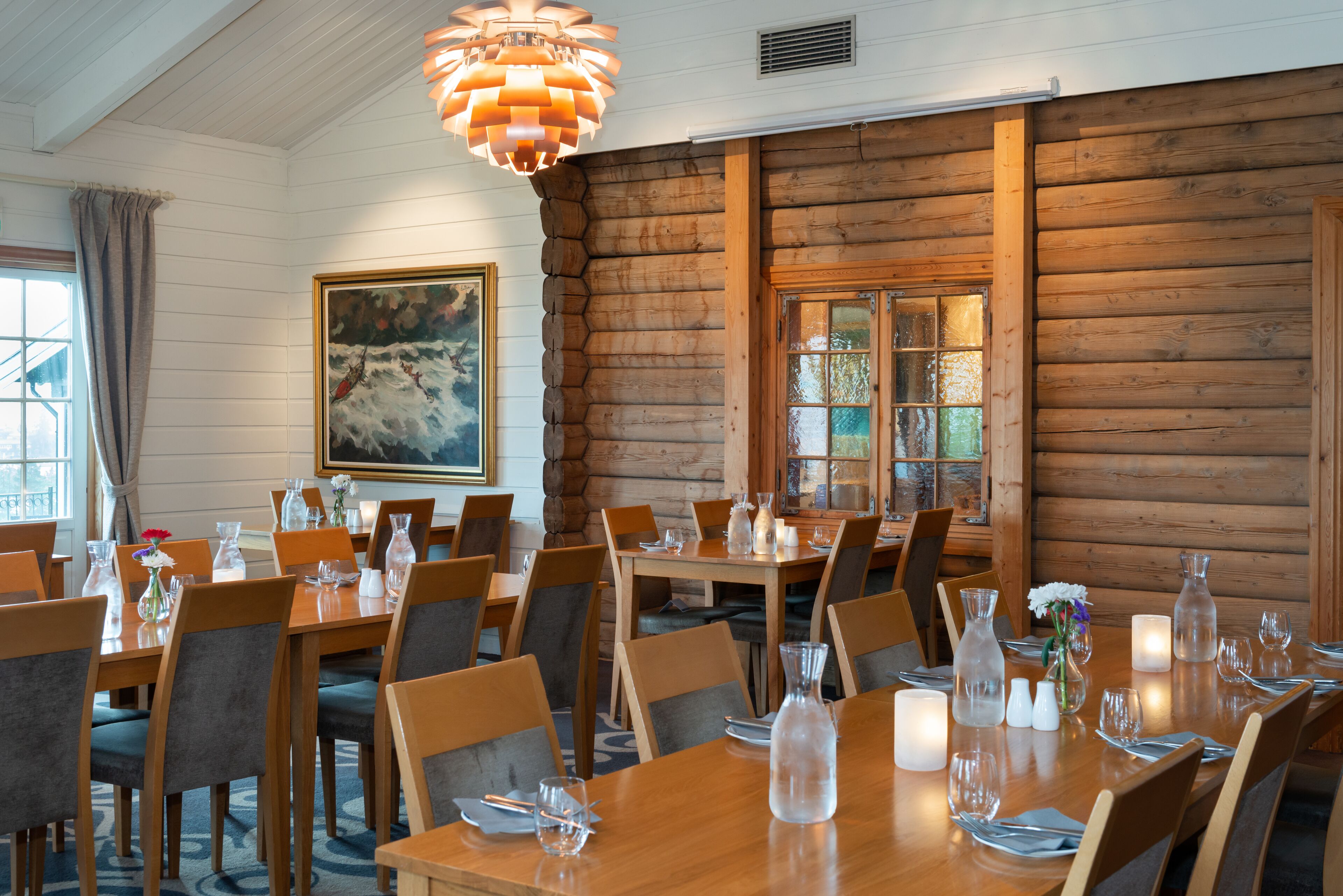A dining area with wooden tables and chairs, candles, and a large painting on the wall.
