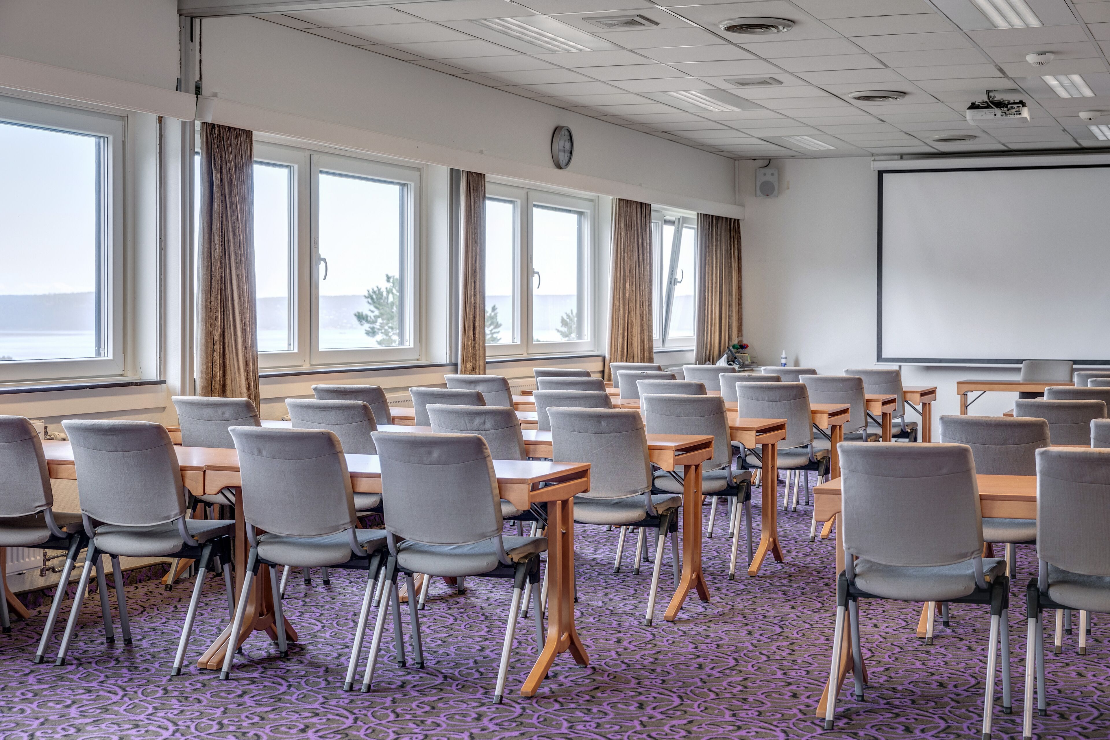 Un salón de conferencias con varias mesas y sillas, ventanas amplias con cortinas, una pantalla de proyector y un reloj en la pared.