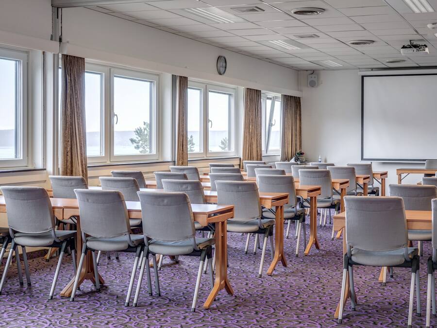 A conference room with multiple tables and chairs, large windows with curtains, a projector screen, and a clock on the wall.