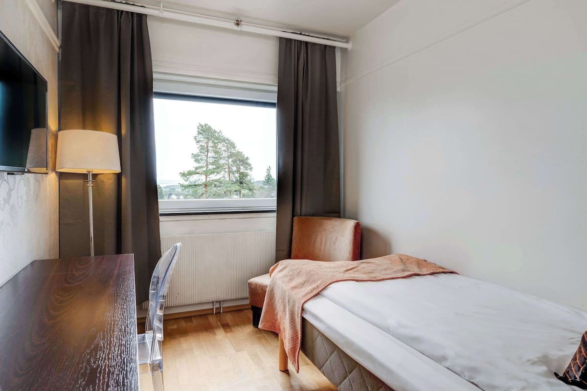 A hotel room with a bed, a desk, a chair, a lamp, and a window with curtains.