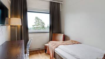 Una habitación de hotel con cama, escritorio, silla, lámpara y ventana con cortinas.