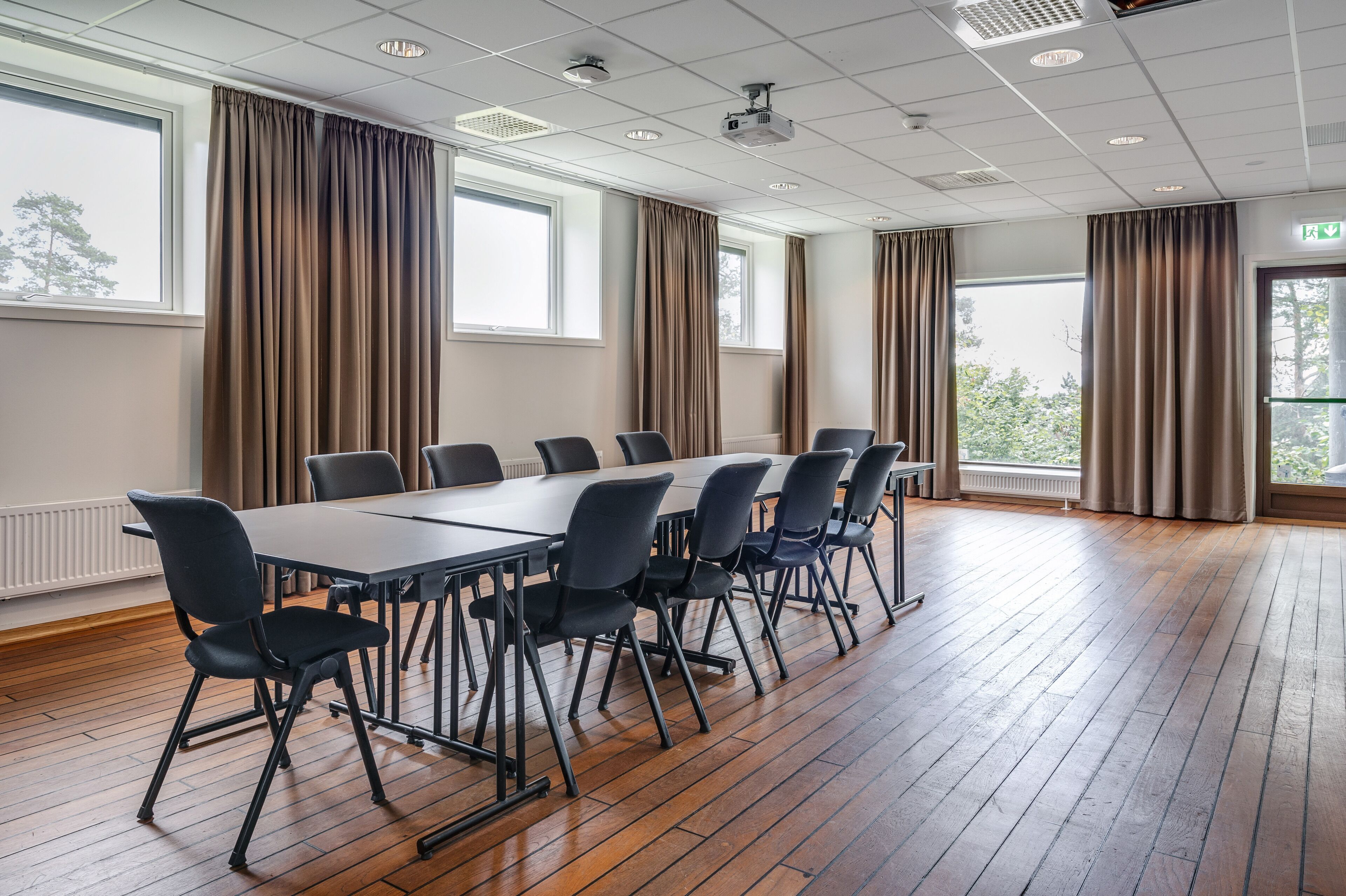 Een congreszaal met een lange tafel en stoelen, grote ramen met gordijnen, houten vloeren en plafondlampen.