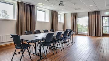 Een congreszaal met een lange tafel en stoelen, grote ramen met gordijnen, houten vloeren en plafondlampen.