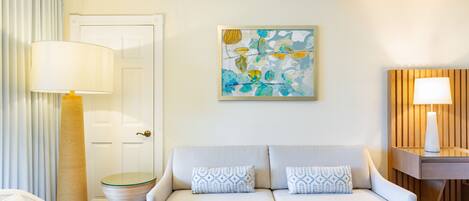 A modern living room with a sofa, a coffee table with a plant, a glass-topped side table, and a painting on the wall.