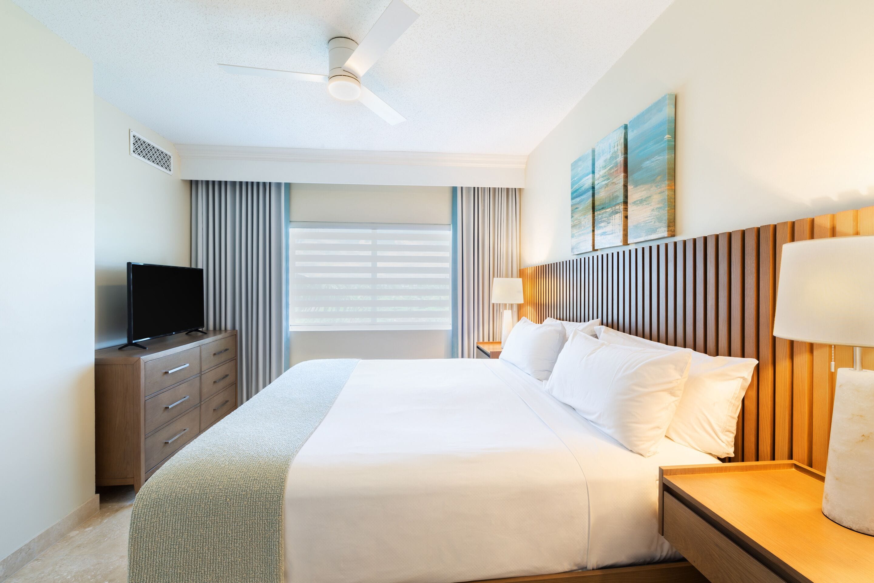 A hotel room with a bed, a television on a wooden dresser, a ceiling fan, and a window with blinds.