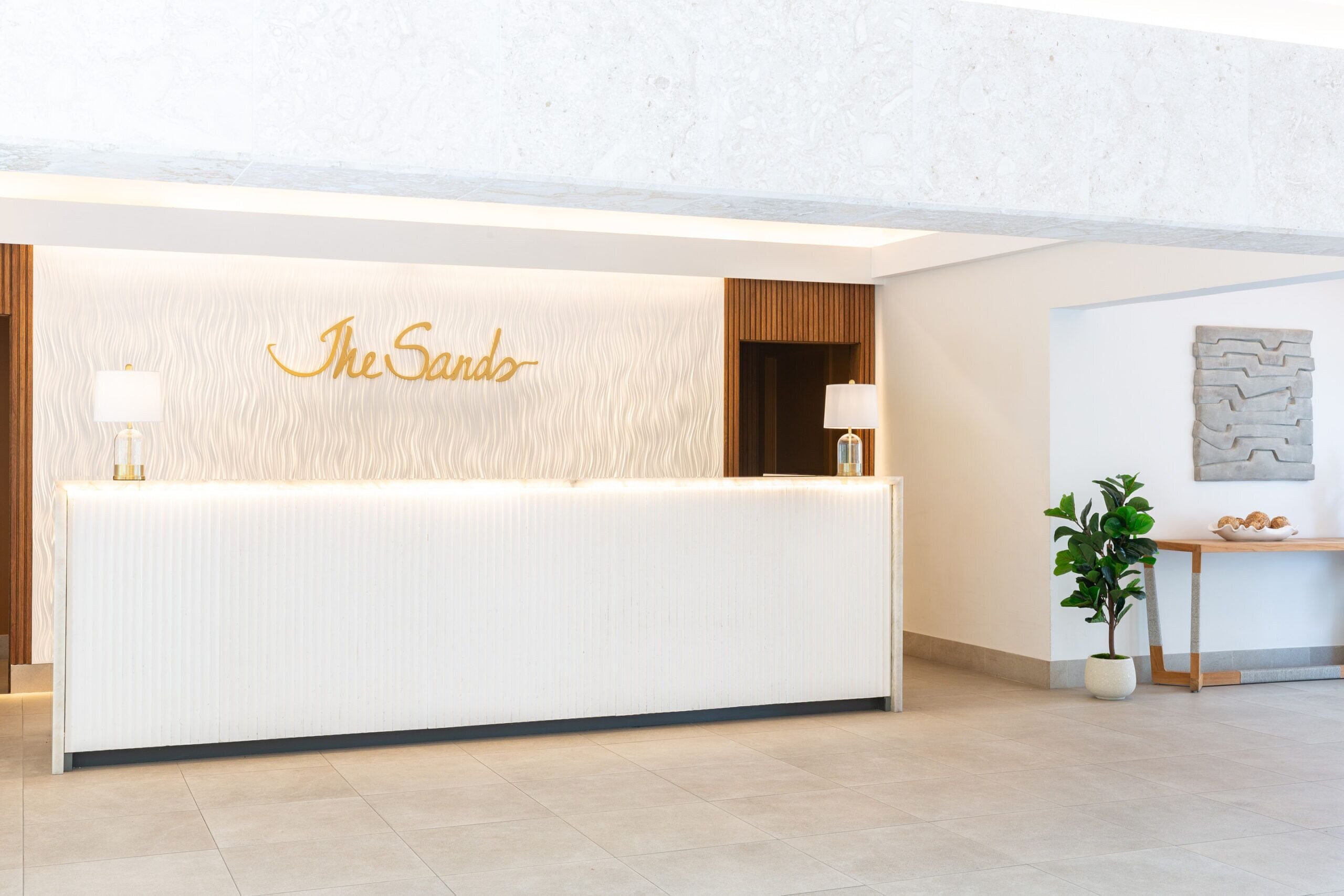 A reception desk with a 'The Sands' sign, two lamps, a plant, and a wall art piece.