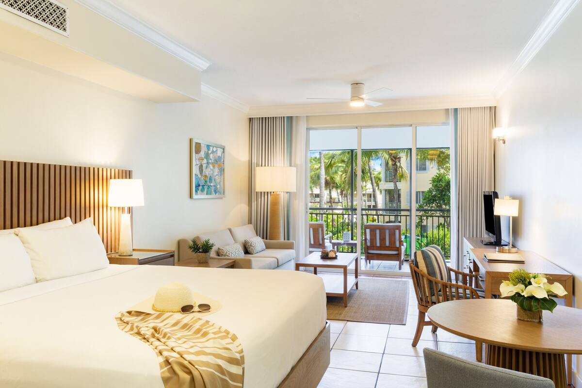 A hotel room with a large bed, a desk, a chair, a small table, and a balcony with a view of palm trees.