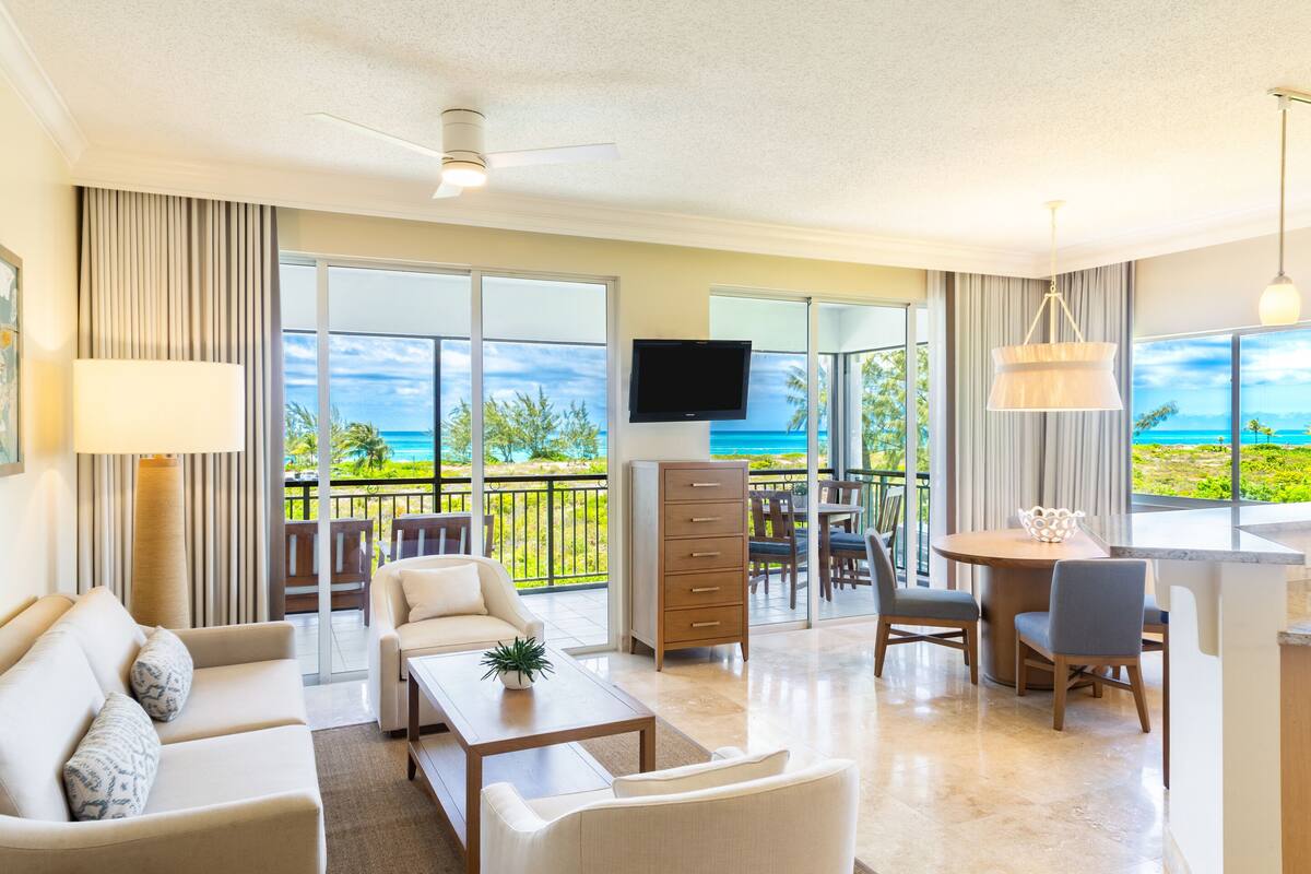 A spacious living room with a sofa, coffee table, and TV, a dining area with chairs and a table, and a balcony with a view of the ocean.