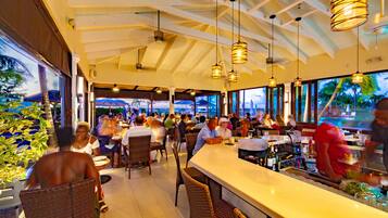 An indoor dining area with wicker chairs, a bar counter, and large windows offering a view of palm trees.