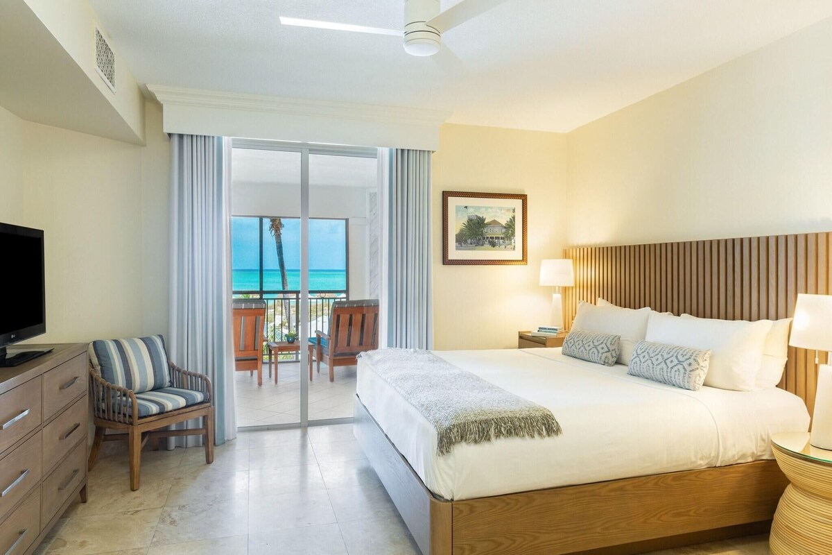 A hotel room with a bed, a television, a chair, a balcony with a view of the ocean, and a painting on the wall.