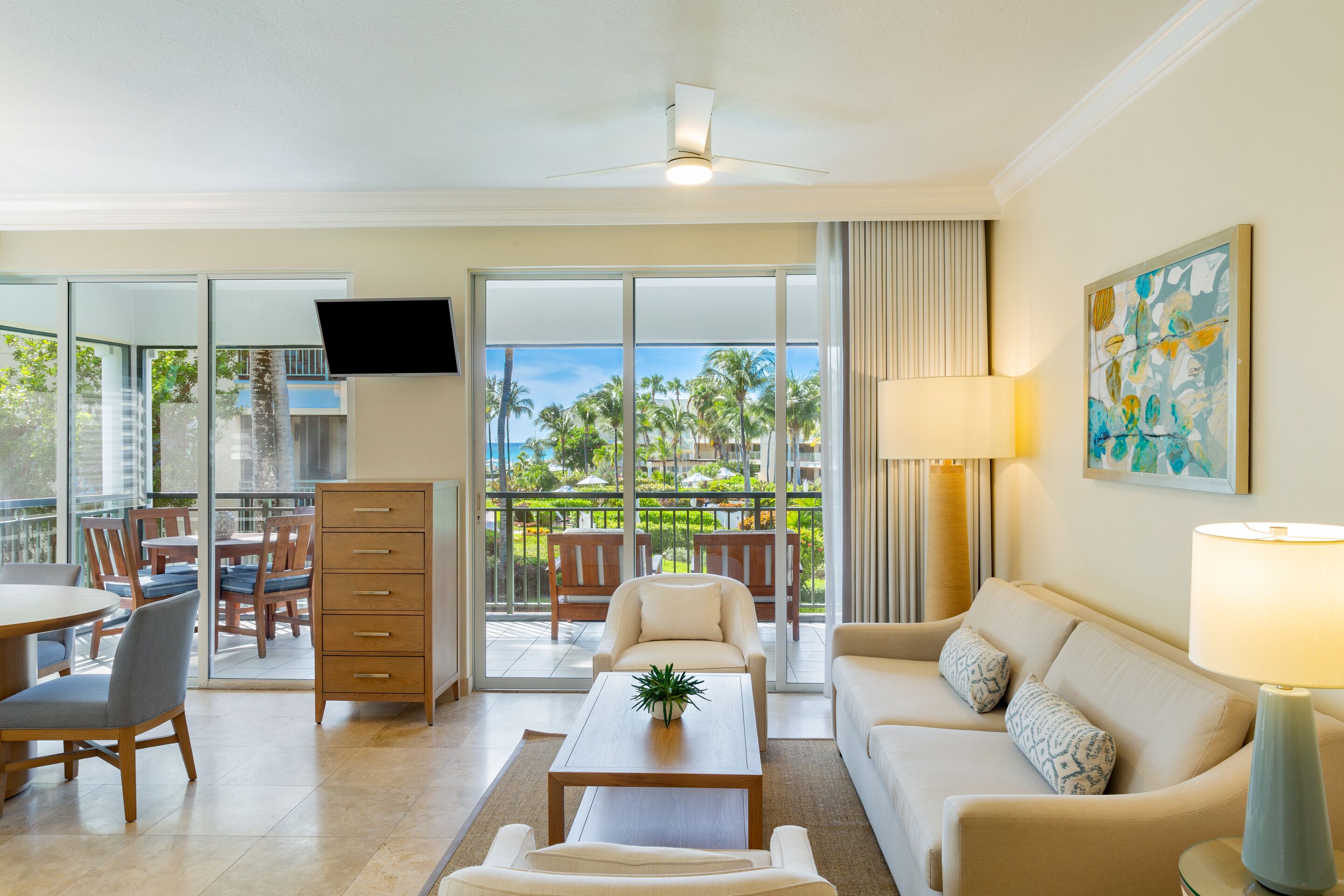 A modern living room with a sofa, coffee table, and TV. There is a balcony with a view of palm trees and a pool.