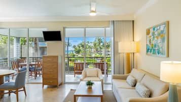 Una sala de estar moderna con sofá, mesa de centro y televisor. Tiene un balcón con vista a palmeras y una piscina.
