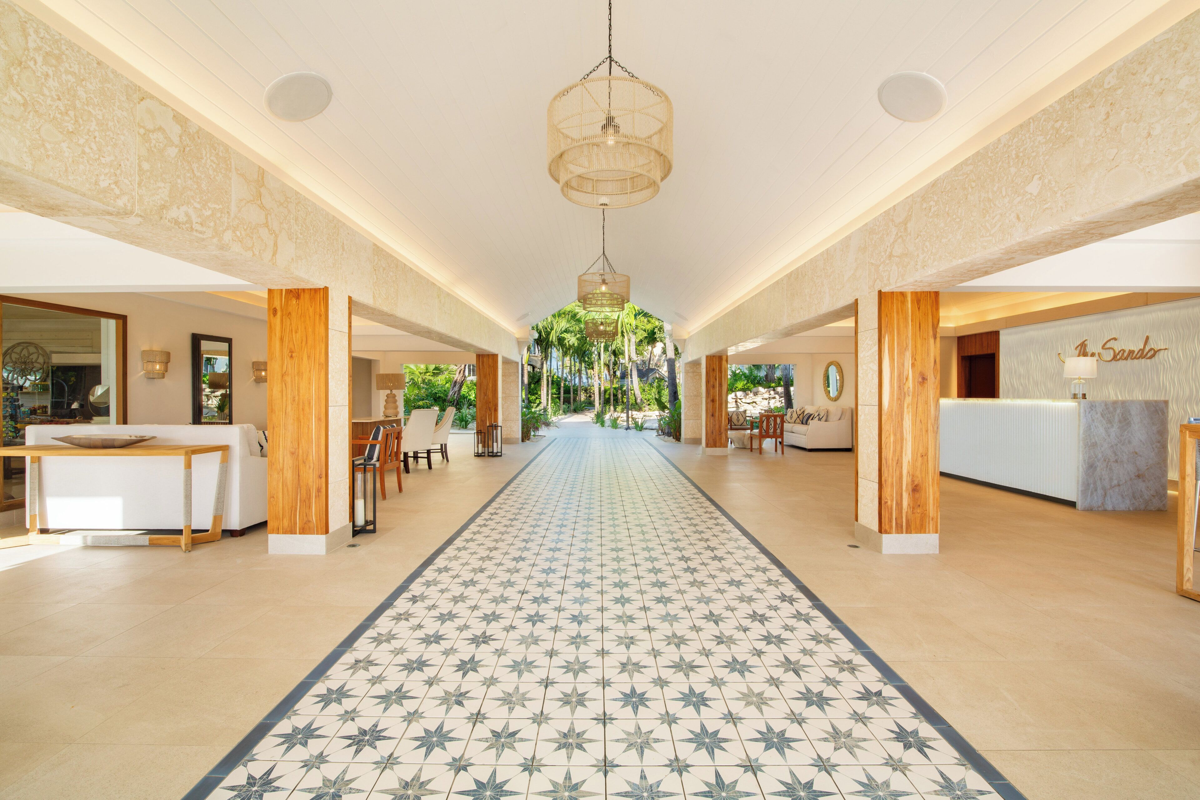 Un hall d’hôtel avec un long couloir carrelé, des piliers en bois, un comptoir d’accueil et un luminaire suspendu.