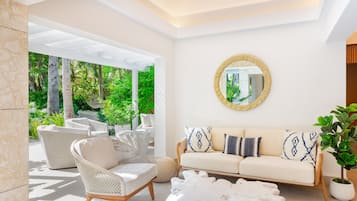 Un salon moderne avec un canapé, des fauteuils et une table basse. Un grand miroir orne le mur, et de l’extérieur, on peut admirer la verdure.