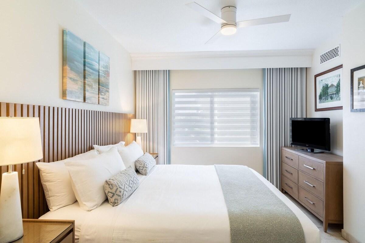 A hotel room with a bed, bedside lamps, a television on a wooden cabinet, a ceiling fan, and a window with blinds.