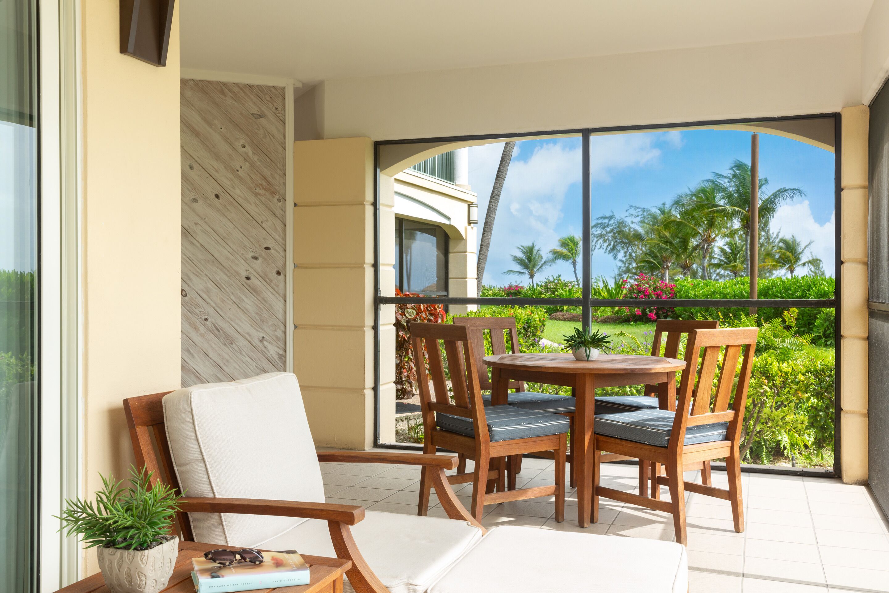 A balcony with a wooden table and chairs, a potted plant, and a view of a building and greenery.
