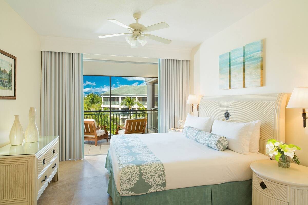 A hotel room with a bed, a ceiling fan, a balcony with a chair, and a view of a building.