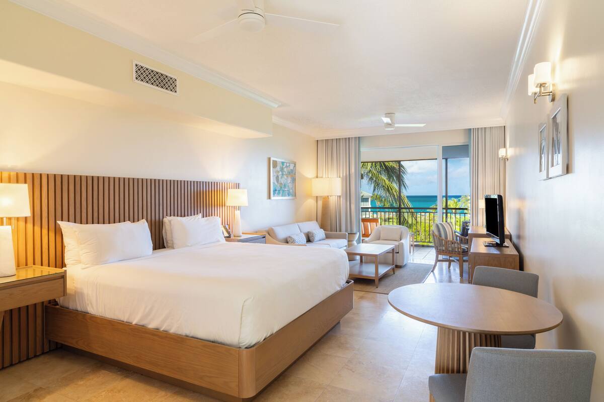 A hotel room with a large bed, a desk, a chair, a round table, and a balcony with a view of the ocean.