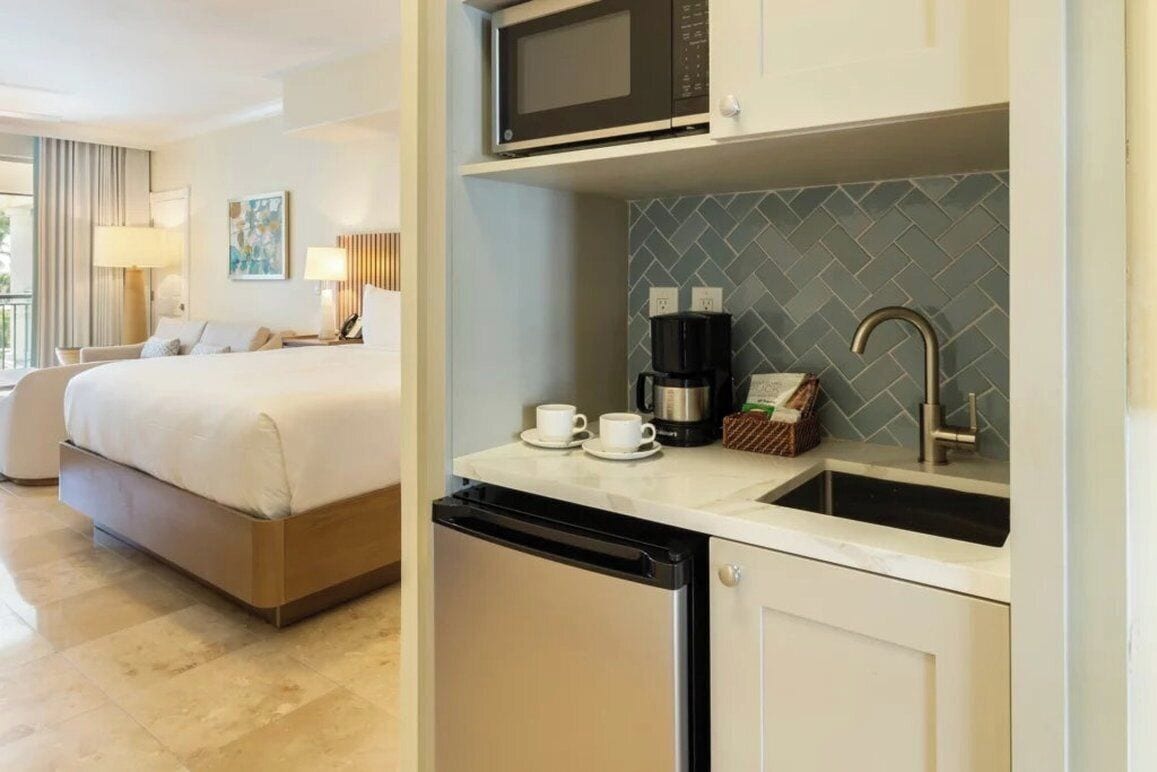 A hotel room with a bed, a small kitchenette, a microwave, a coffee maker, and a sink.