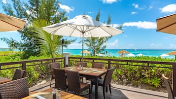 A wooden deck with wicker chairs and tables, overlooking a beach with umbrellas and a clear blue sky.