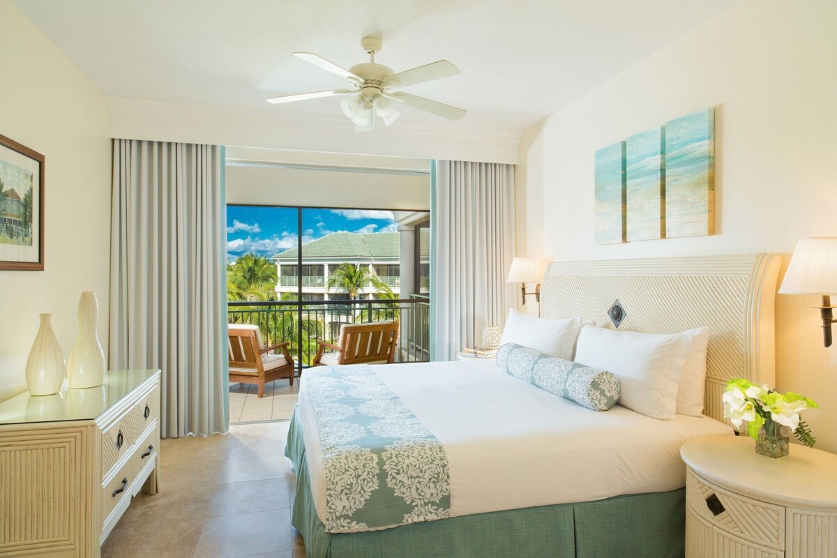 A hotel room with a bed, a nightstand, a dresser, a ceiling fan, a balcony with chairs, and a view of a building.