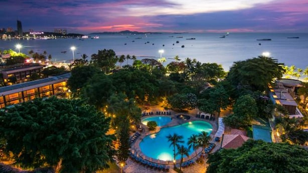 Un resort con una amplia piscina rodeada de palmeras y sillas de descanso, con vista a una masa de agua con barcos.