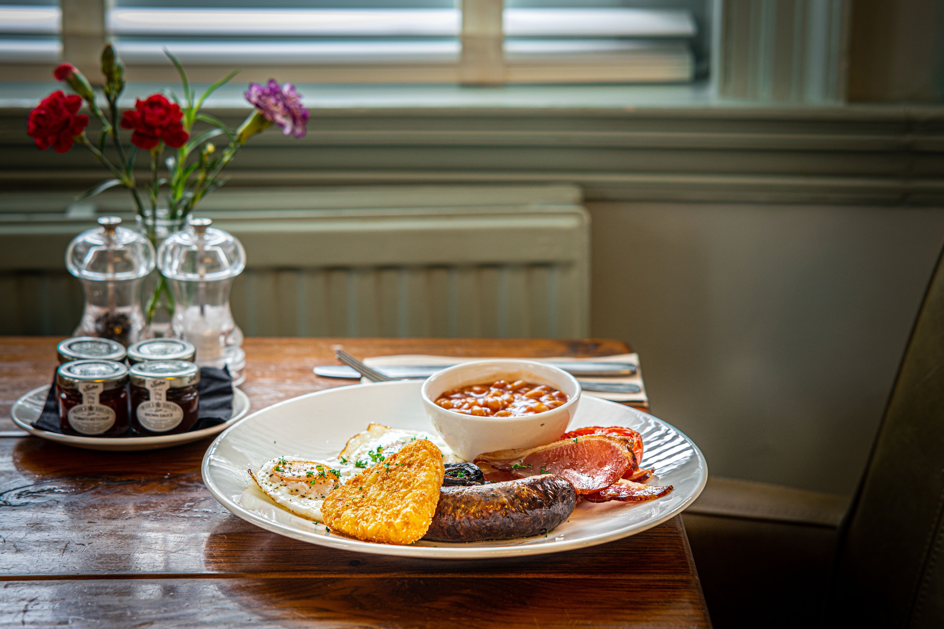 Une assiette de petit-déjeuner avec des œufs, du bacon et des haricots, un pot de confiture et un vase de fleurs sur une table en bois.