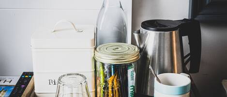 Una caja de madera con la etiqueta '¿Bebidas para alguien?', que contiene una botella de vidrio, un termo, un frasco de bolsitas de té, una tetera y una taza.