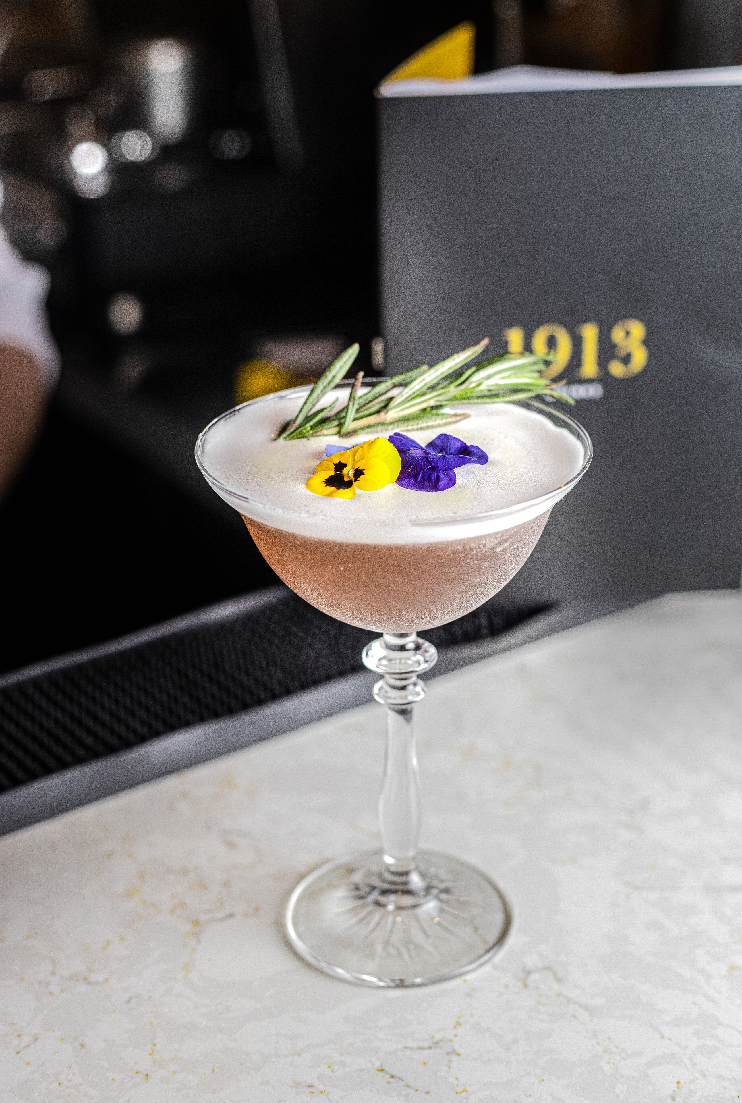 A cocktail in a martini glass with a sprig of rosemary and a flower garnish on a marble surface.