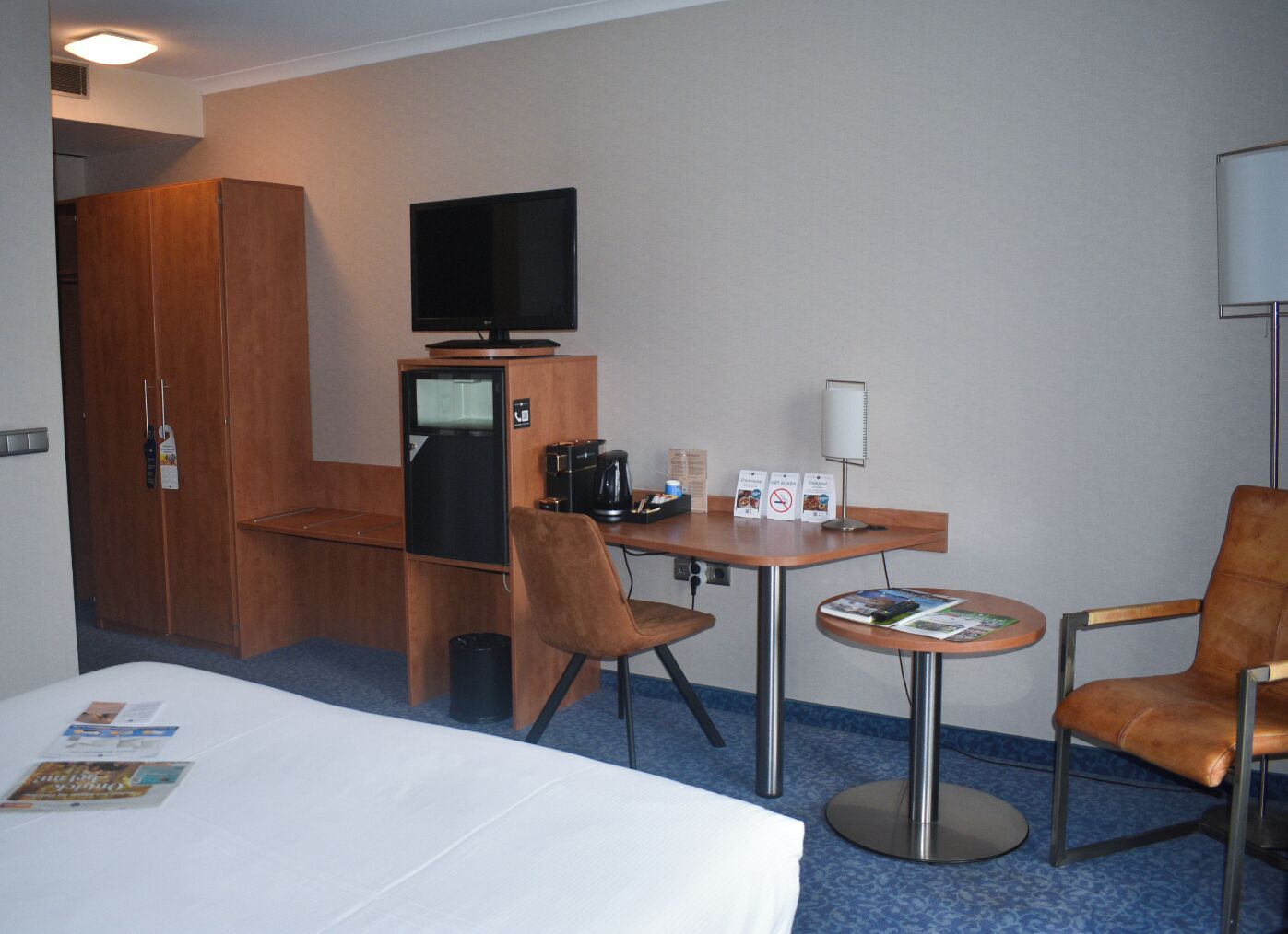 A hotel room with a bed, a desk with a computer, a television, and two chairs.