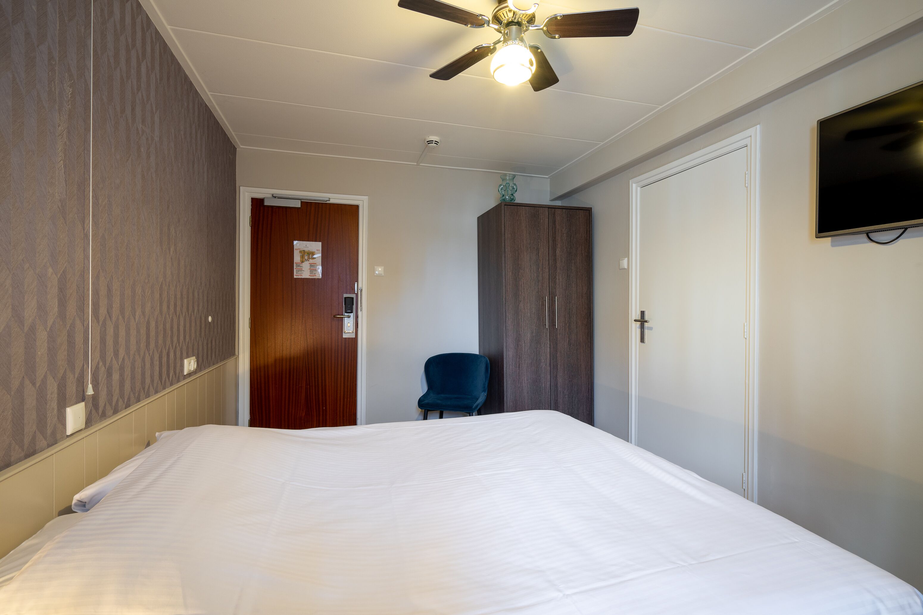 Habitación de hotel con una cama grande, un ventilador de techo, un televisor montado en la pared y una puerta de madera.