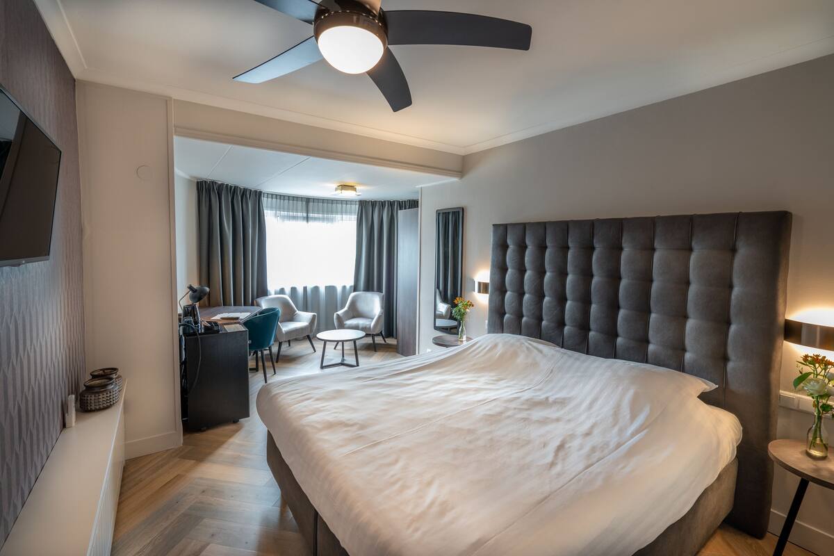 Une chambre d’hôtel moderne dotée d’un grand lit, d’un ventilateur de plafond, d’un bureau avec une chaise et offrant une vue sur l’extérieur par la fenêtre.