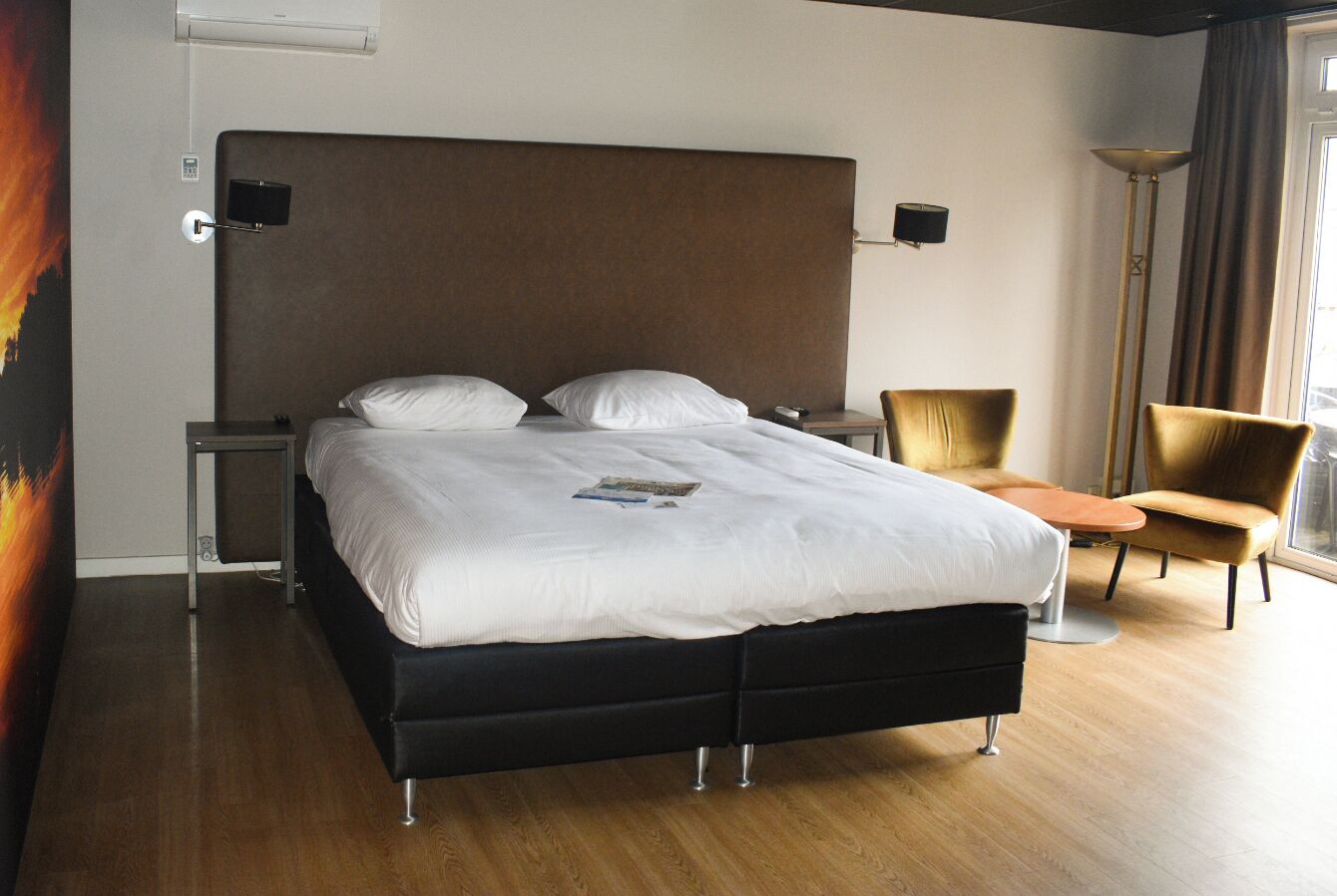 A modern hotel room with a large bed, a bedside table, a chair, and a floor lamp.