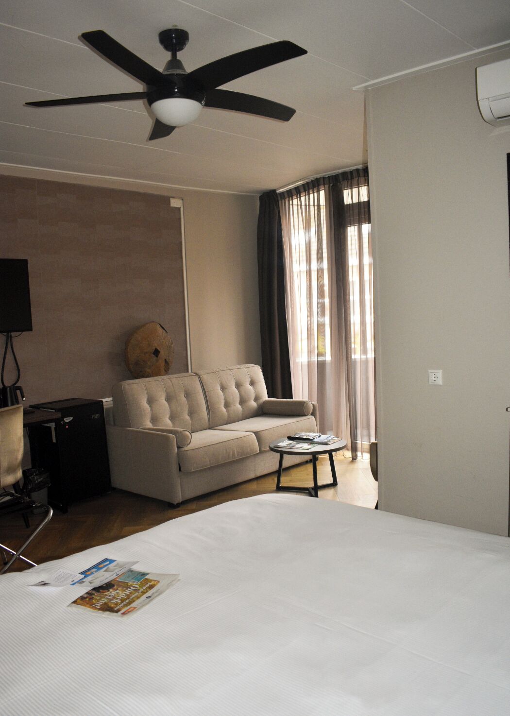 A hotel room with a bed, a sofa, a small table, a ceiling fan, and a window with curtains.