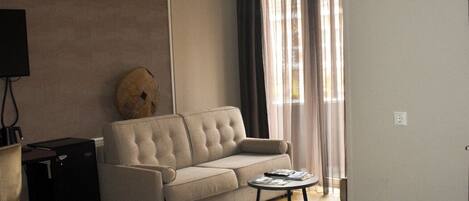 Habitación de hotel con cama, sofá, una mesita, ventilador de techo y ventana con cortinas.