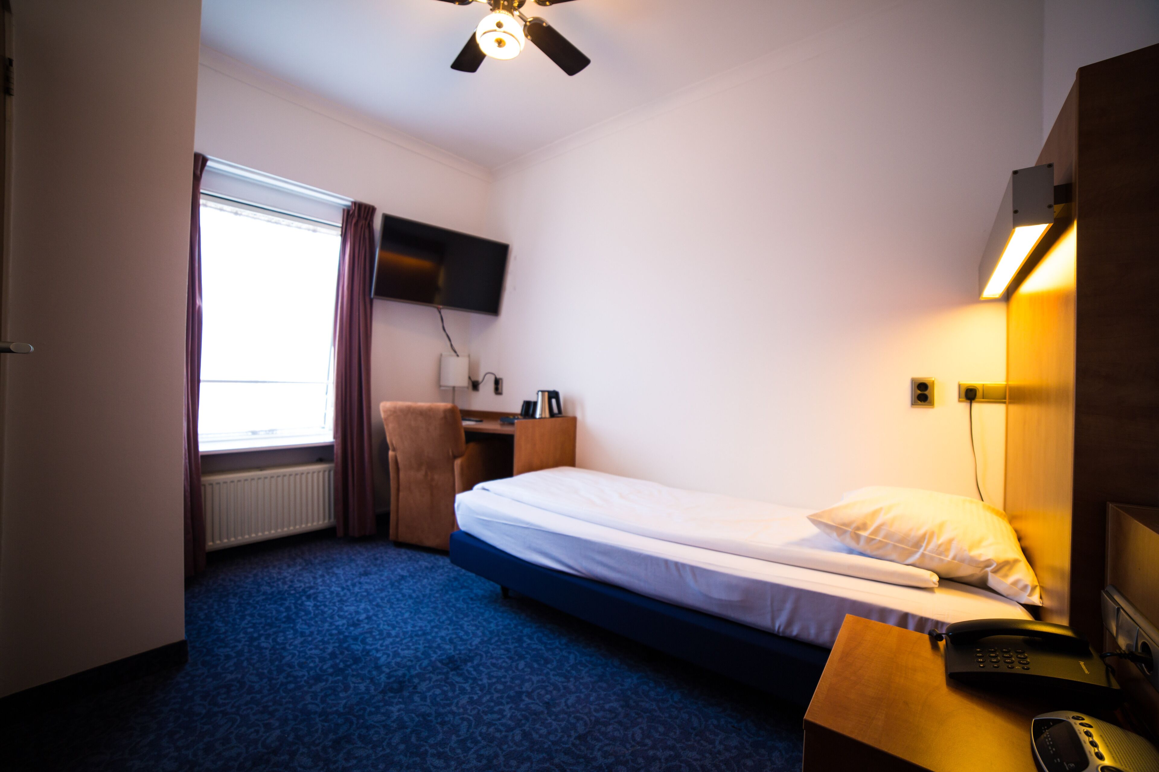 Una habitación de hotel con una cama individual, un escritorio con silla, un televisor fijado a la pared y un ventilador de techo.